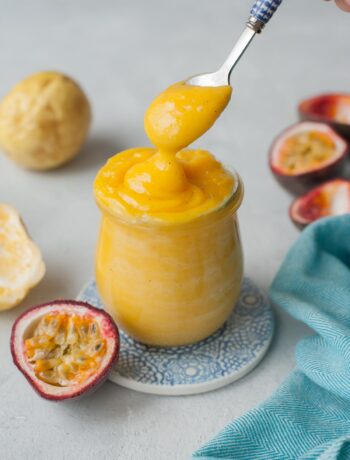 Passion fruit curd is being spooned into a jar. Passion fruits and a blue kitchen cloth in the background.