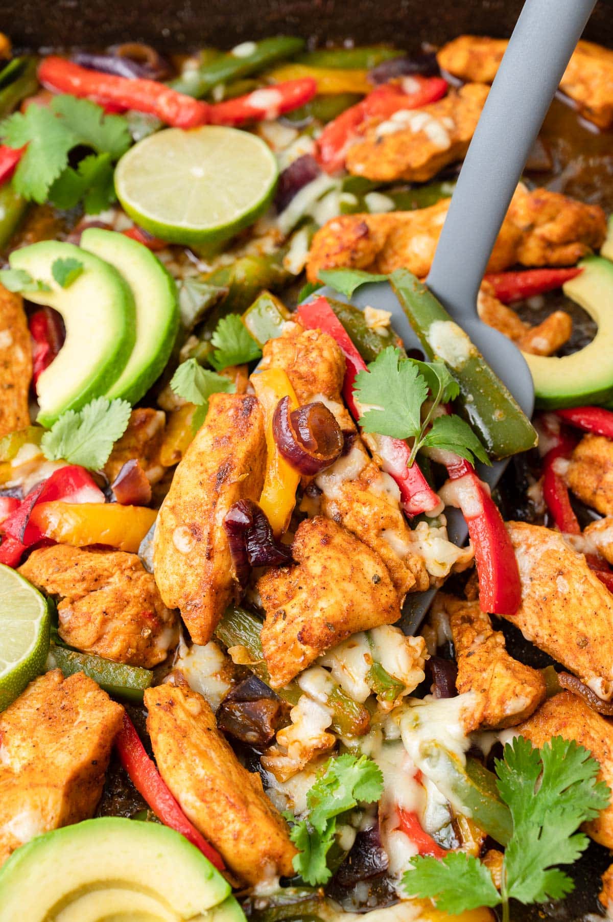 Sheet pan chicken fajitas on a black baking sheet. A grey spatula is picking up portion of the dish.