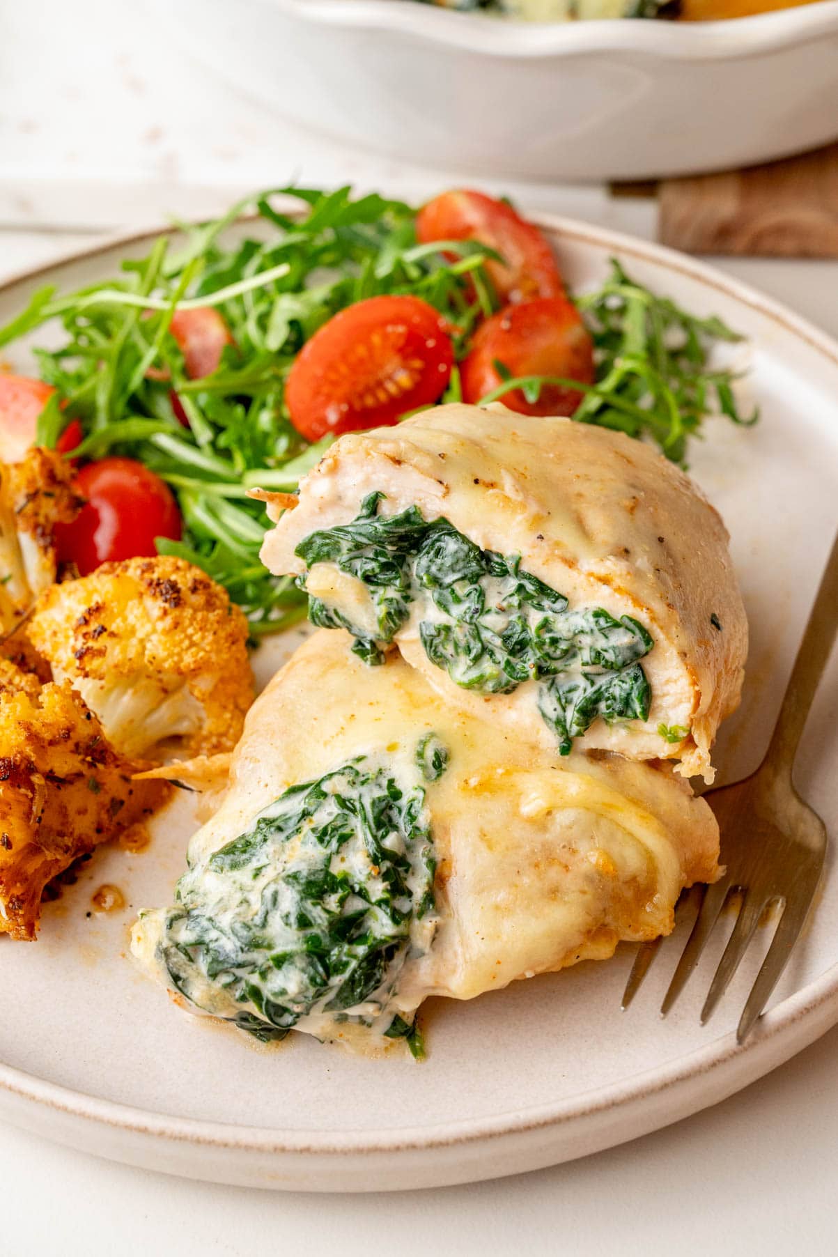 A spinach-stuffed chicken breast sliced in half, revealing a creamy spinach and cheese filling. The dish is topped with melted cheese and served with a side of arugula salad with cherry tomatoes and roasted cauliflower.