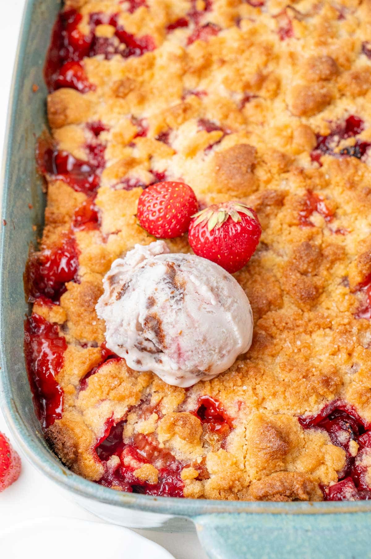 Baked strawberry rhubarb crumble in a baking dish, topped with fresh strawberries and a scoop of melting ice cream.

