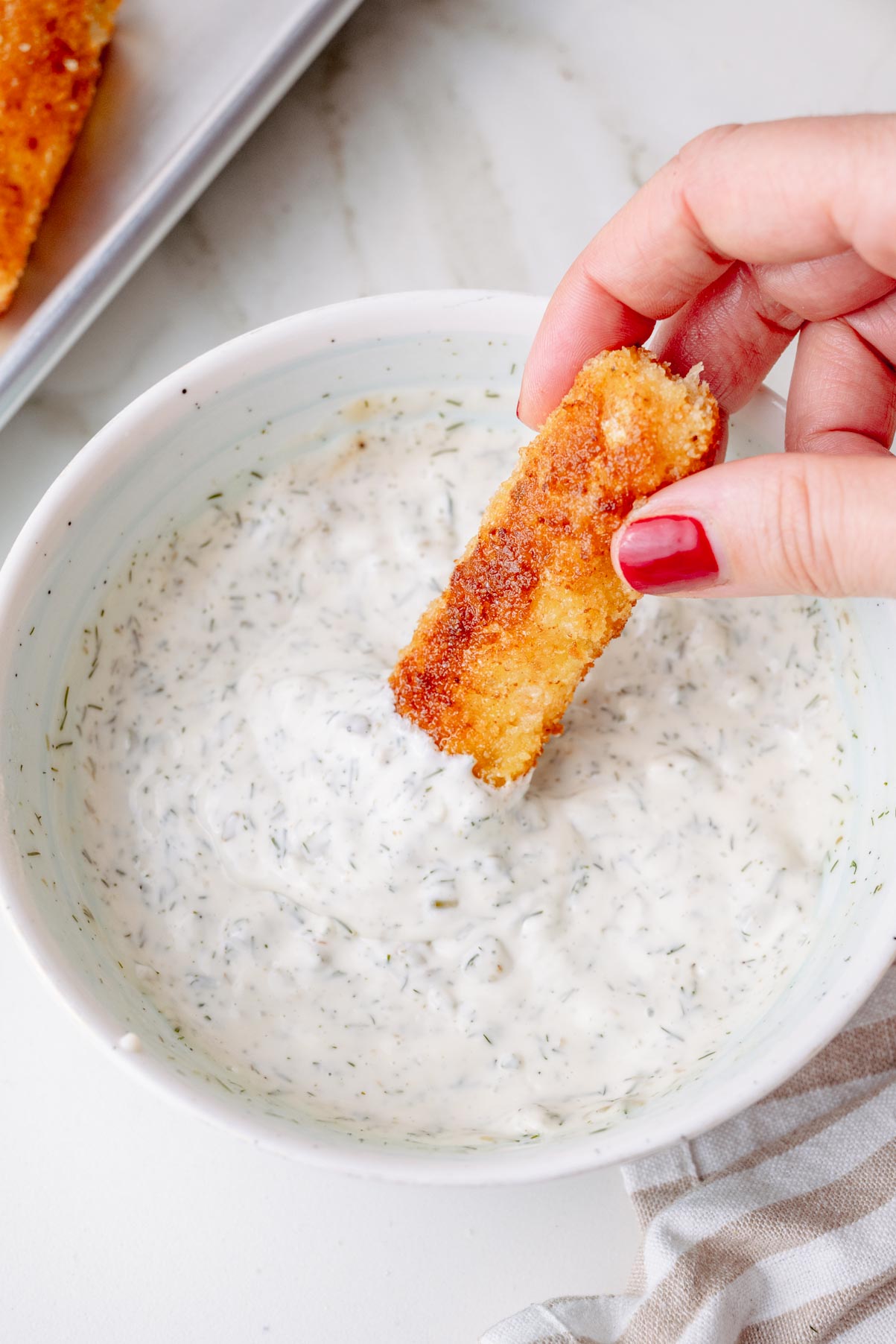 A hand dipping a crispy, golden fish stick into a bowl of tartar sauce. The sauce is speckled with chopped dill and pickles, and the fish sticks are arranged on a tray nearby.