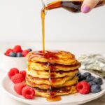 A stack of yogurt pancakes on a plate with barries. Maple syrup is being poured over.