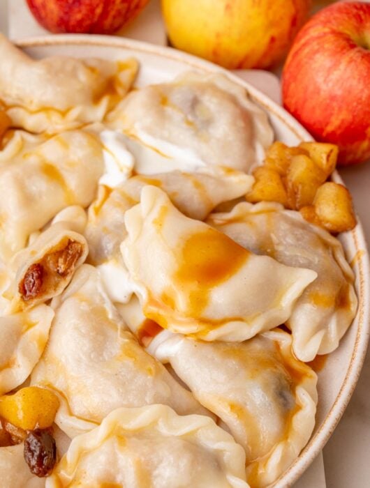 Apple pierogi on a beige plate served with caramel sauce and sour cream.