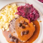 A slice of Sauerbraten roast with a brown sauce and raisins served with Spätzle and red cabbage on a plate.