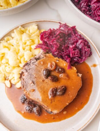 A slice of Sauerbraten roast with a brown sauce and raisins served with Spätzle and red cabbage on a plate.