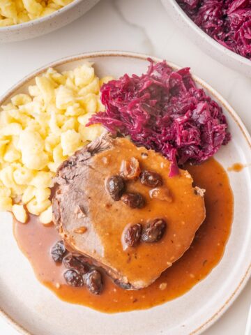 A slice of Sauerbraten roast with a brown sauce and raisins served with Spätzle and red cabbage on a plate.